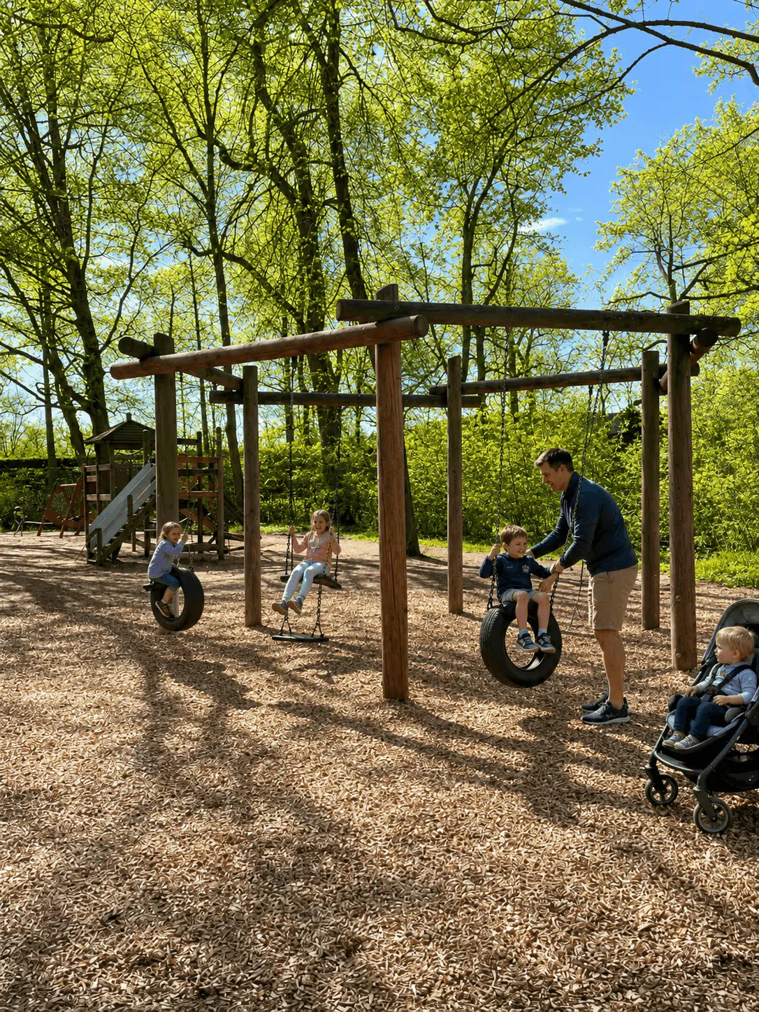 Papa mit Kinderwagen schiebt ein Kind auf der Reifenschaukel auf einem grünen Spielplatz in Würzburg an.